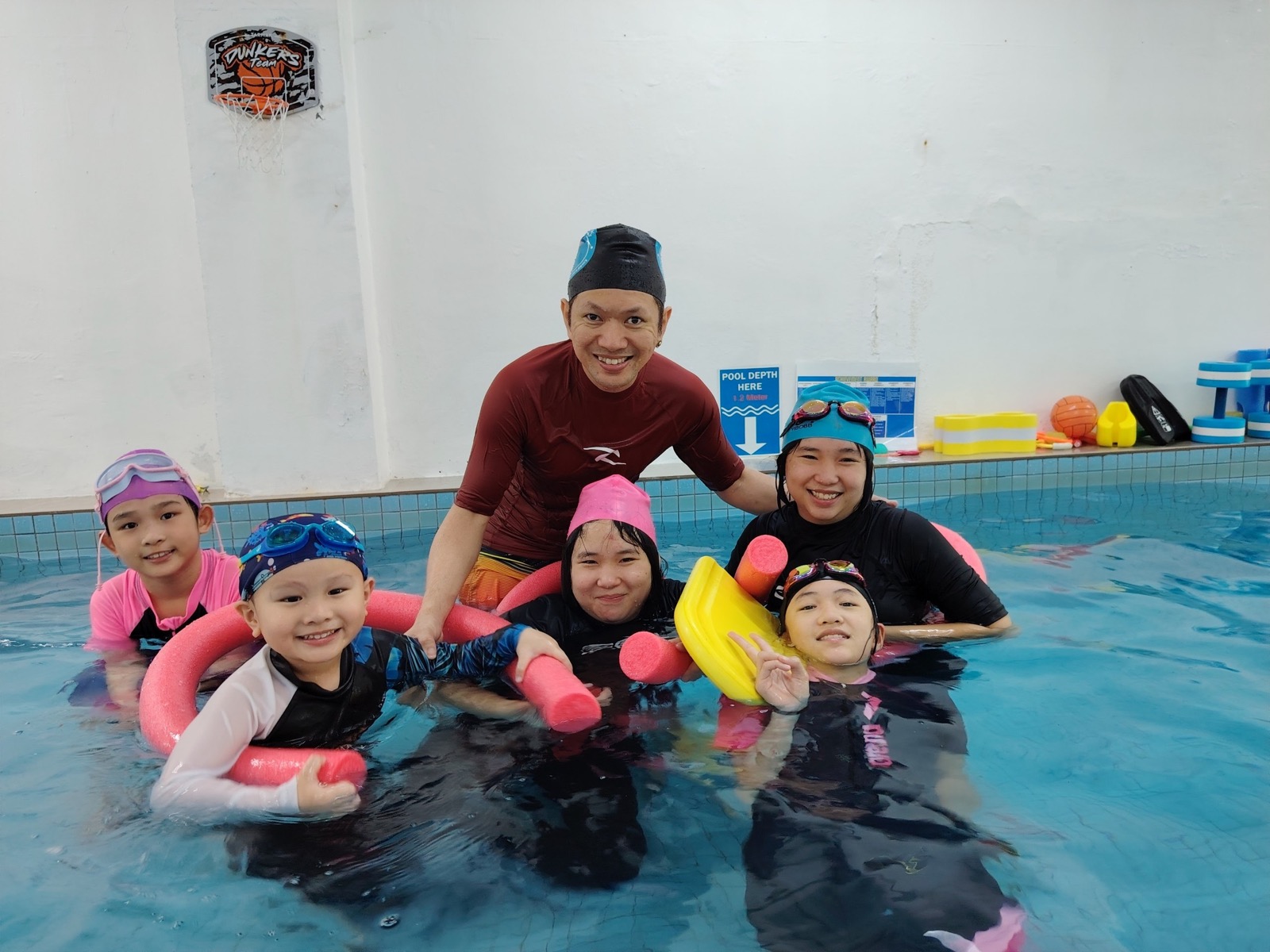 A JC Swimming coach guiding four happy children during a group lesson in the warm indoor pool
